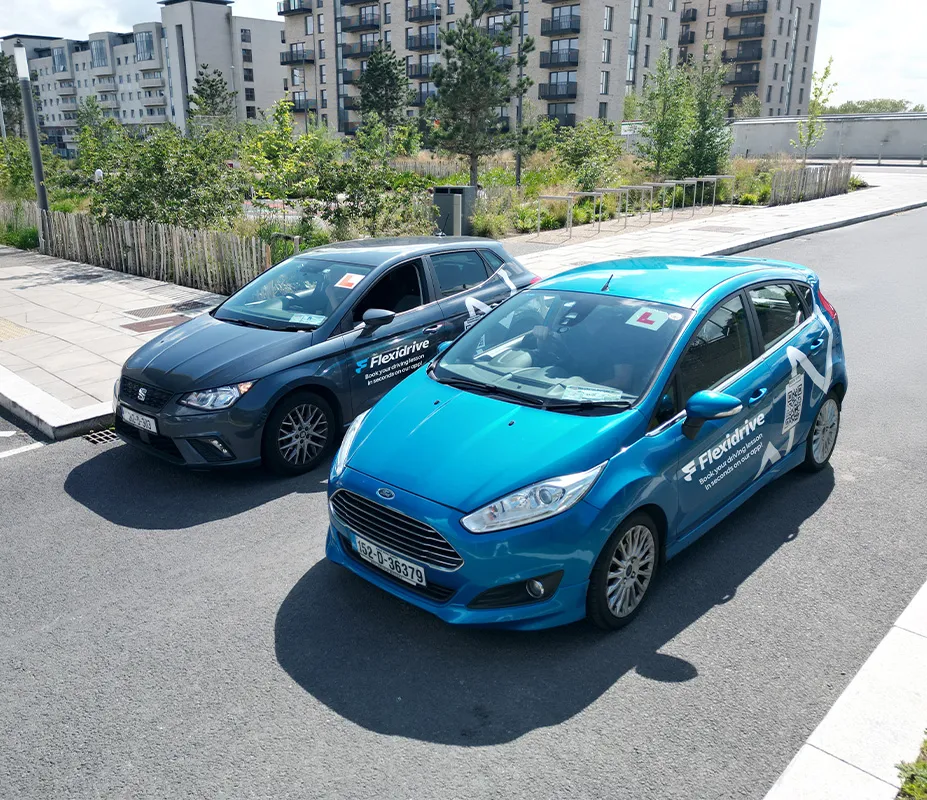 Two parked Flexidrive driving school cars, one blue and one dark gray, with 'L' learner signs on the windshields on a sunny street near modern apartment buildings.