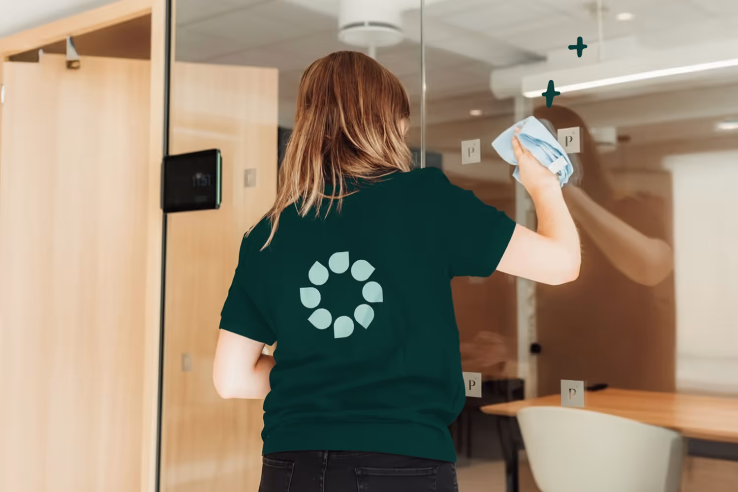 Person med grønn t-skjorte vasker glassvegg inne i et lyst moderne kontorlandskap.
