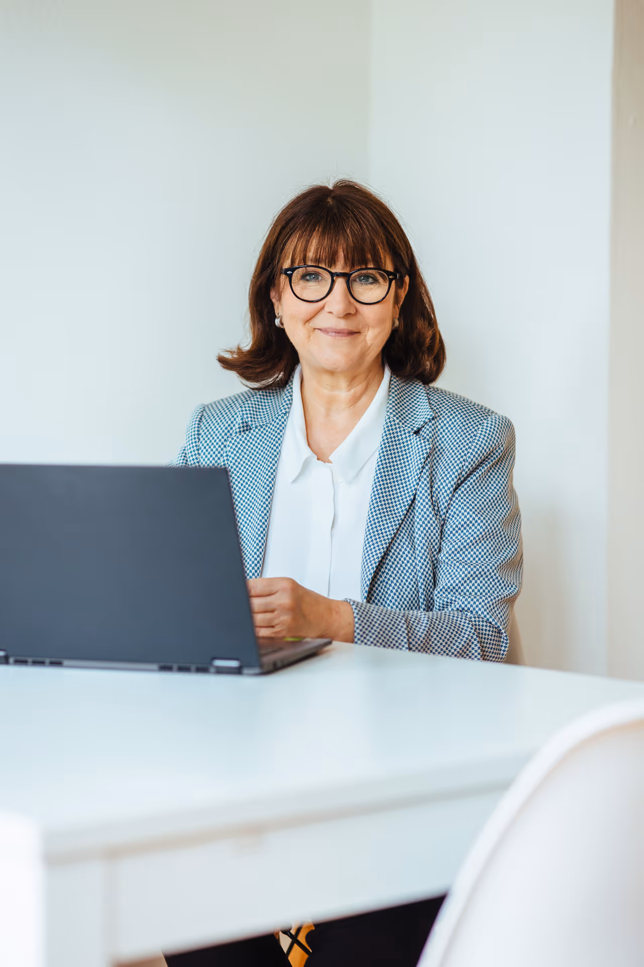 Frau sitzt an einem Schreibtisch mit Laptop, trägt eine Brille und einen hellblauen Blazer – moderne, professionelle Arbeitsumgebung.