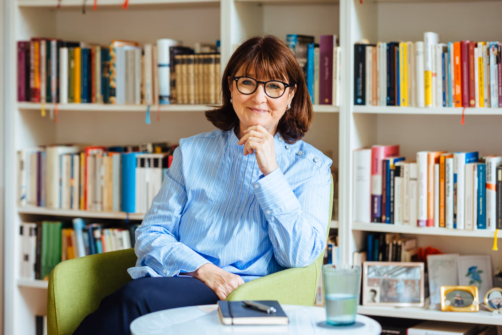 Frau mit Brille sitzt entspannt in einem grünen Sessel vor einem Bücherregal – inspirierende Arbeits- oder Beratungssituation.