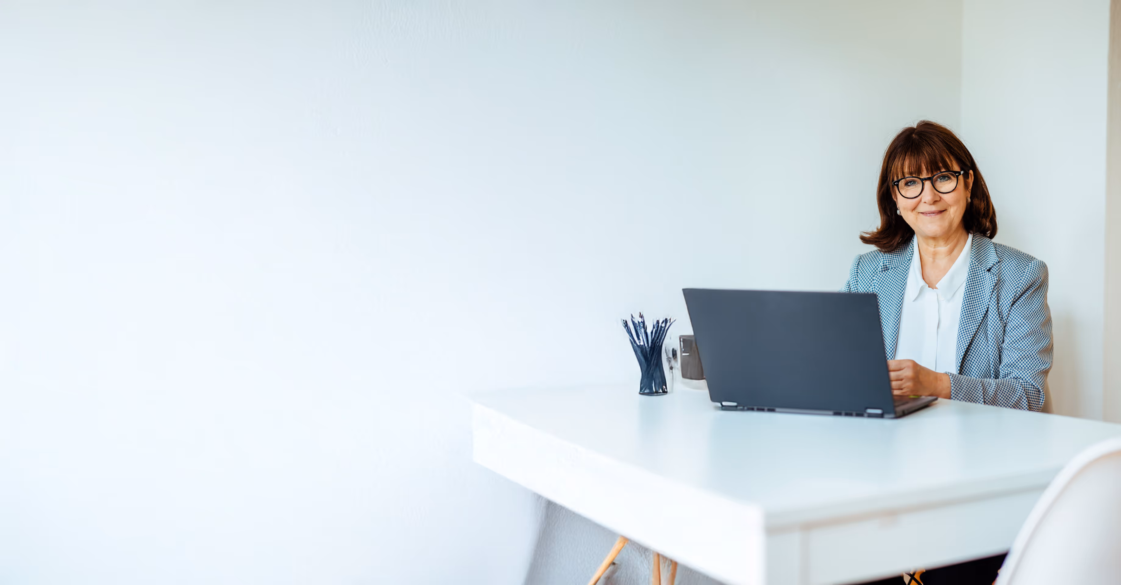 Frau mit Brille sitzt an einem weißen Schreibtisch mit Laptop, lächelt in die Kamera – moderne, minimalistische Büroszene.