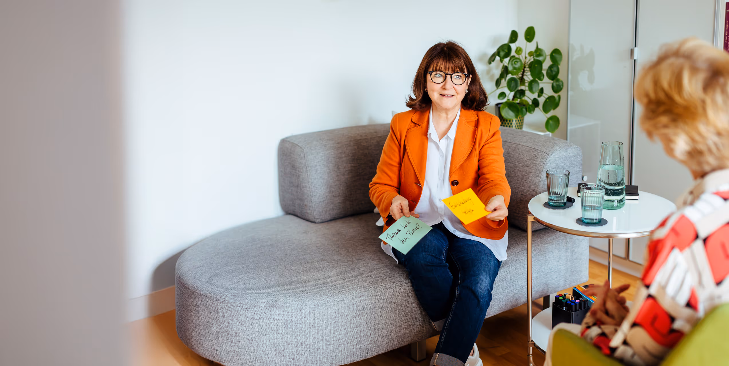 Frau in orangefarbener Jacke sitzt auf einem Sofa, zeigt bunte Moderationskarten, im Gespräch mit einer weiteren Frau – Coaching-Situation in heller Praxisumgebung.