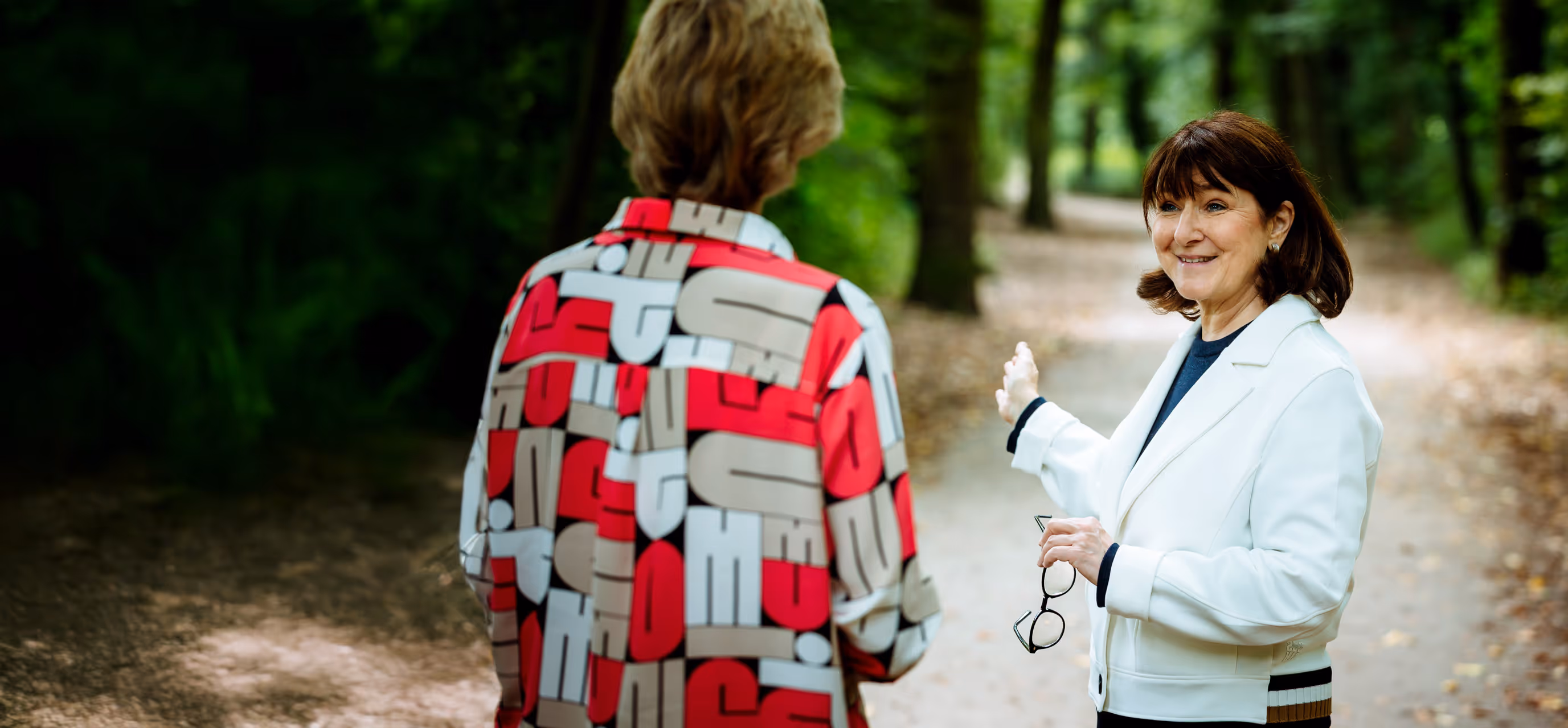 Zwei Frauen im Gespräch auf einem Waldweg, eine zeigt gestikulierend in eine Richtung – freundlicher Austausch in natürlicher Umgebung.