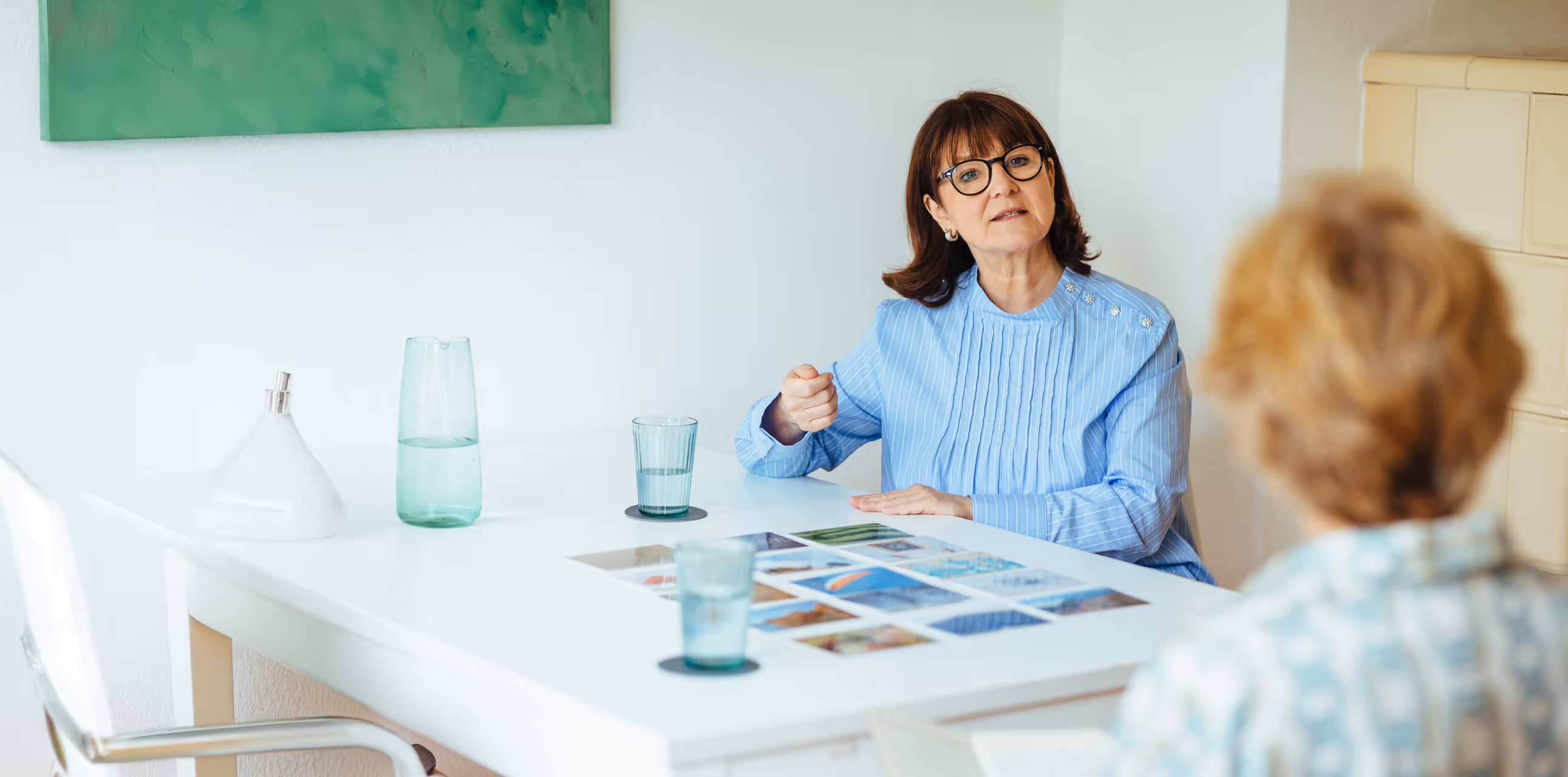 Frau mit Brille erklärt Farbkarten am Tisch in einem Gespräch mit einer anderen Frau – Beratungssituation in heller, ruhiger Umgebung.