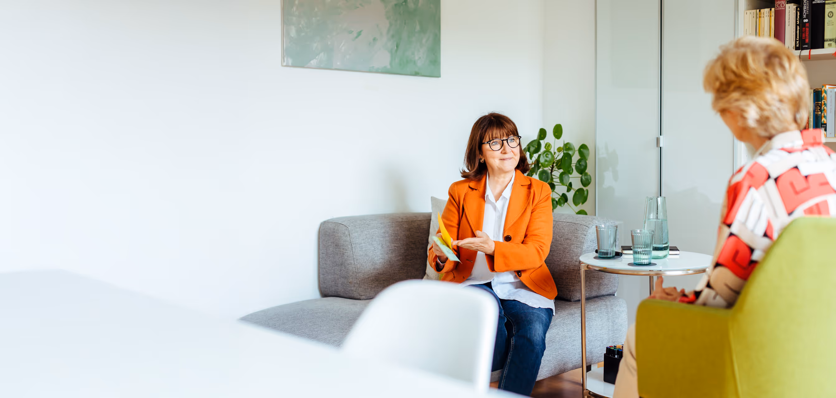 Frau in orangem Blazer spricht mit einer anderen Frau in einem modernen Beratungsraum – Gesprächssituation in professioneller, entspannter Atmosphäre