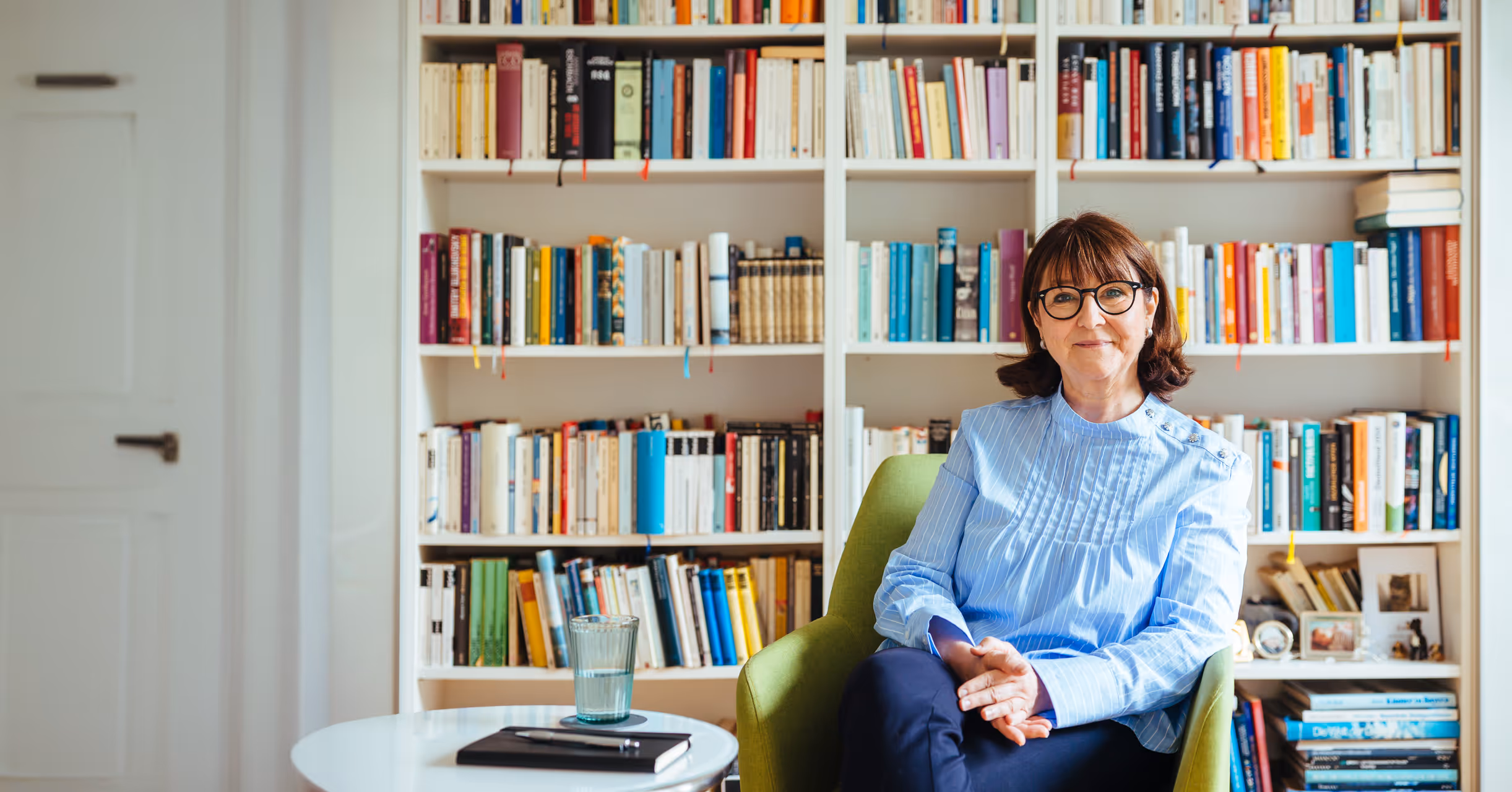 Frau mit Brille sitzt in einem grünen Sessel vor einem großen Bücherregal – ruhige, professionelle Atmosphäre für Beratung oder Coaching
