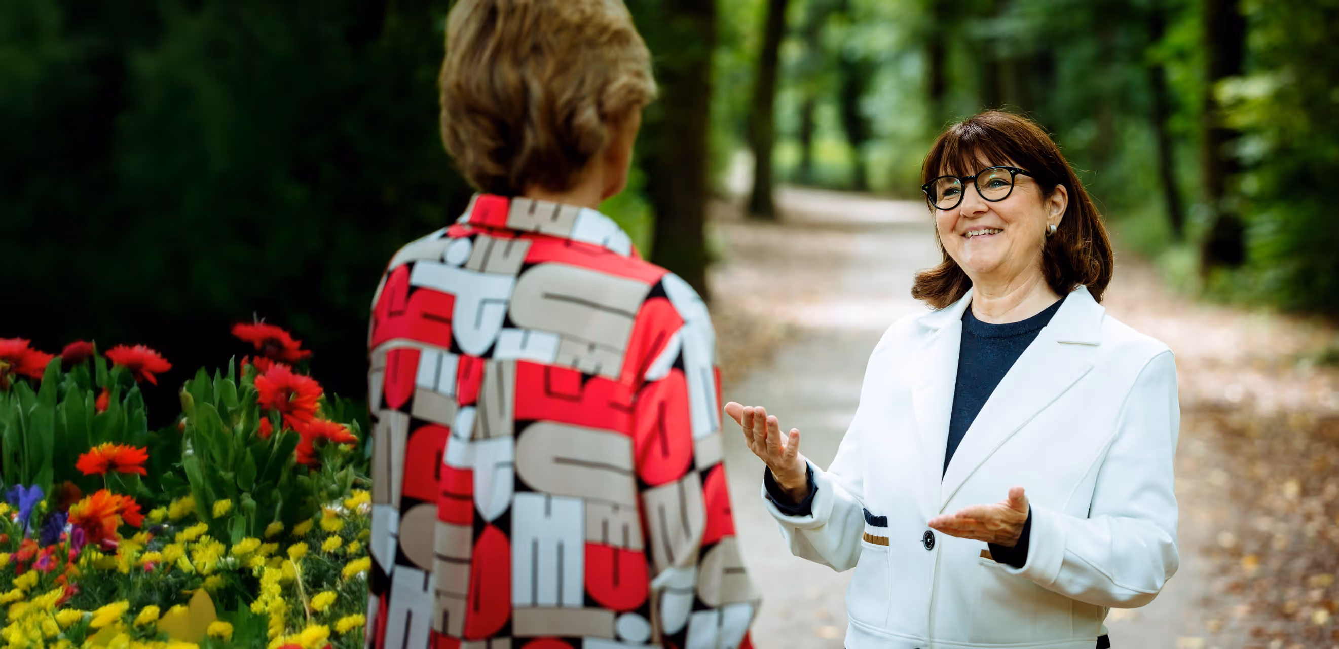 Zwei Frauen im Gespräch auf einem Waldweg, eine von ihnen gestikuliert freundlich – Begegnung in natürlicher Umgebung mit Blumen im Vordergrund.
