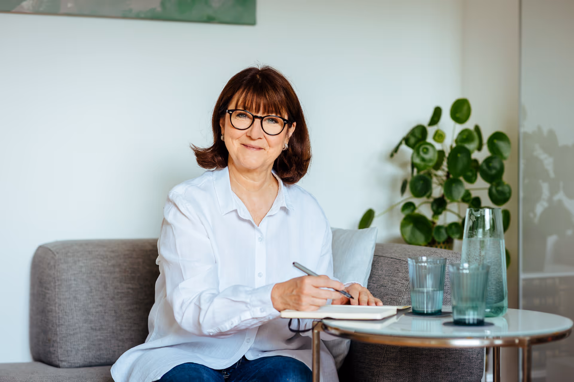 Frau mit Brille sitzt auf einem Sofa, schreibt in ein Notizbuch – ruhige, freundliche Atmosphäre mit Pflanzen im Hintergrund