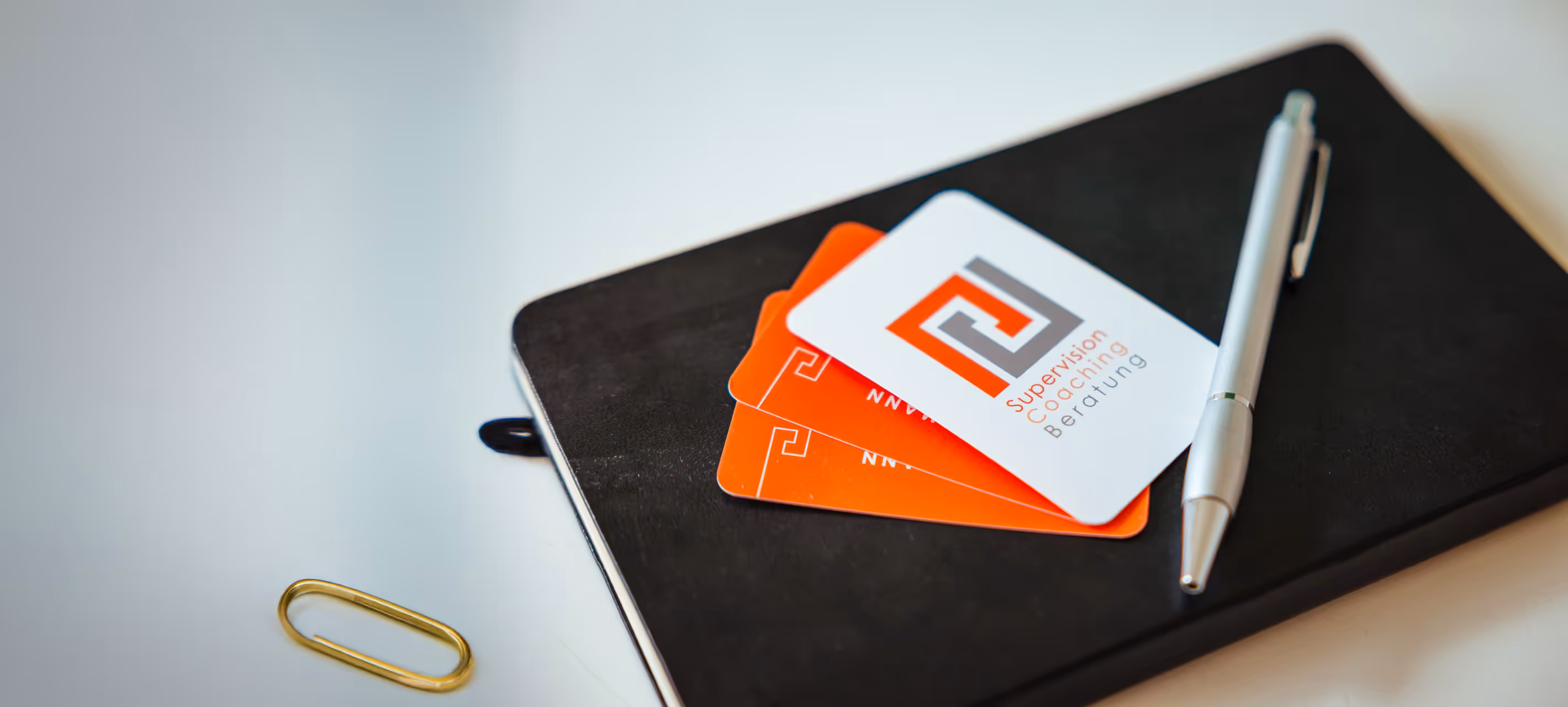 Visitenkarten mit dem Logo und der Aufschrift ‚Supervision, Coaching, Beratung‘ auf einem schwarzen Notizbuch mit Kugelschreiber und Büroklammer.