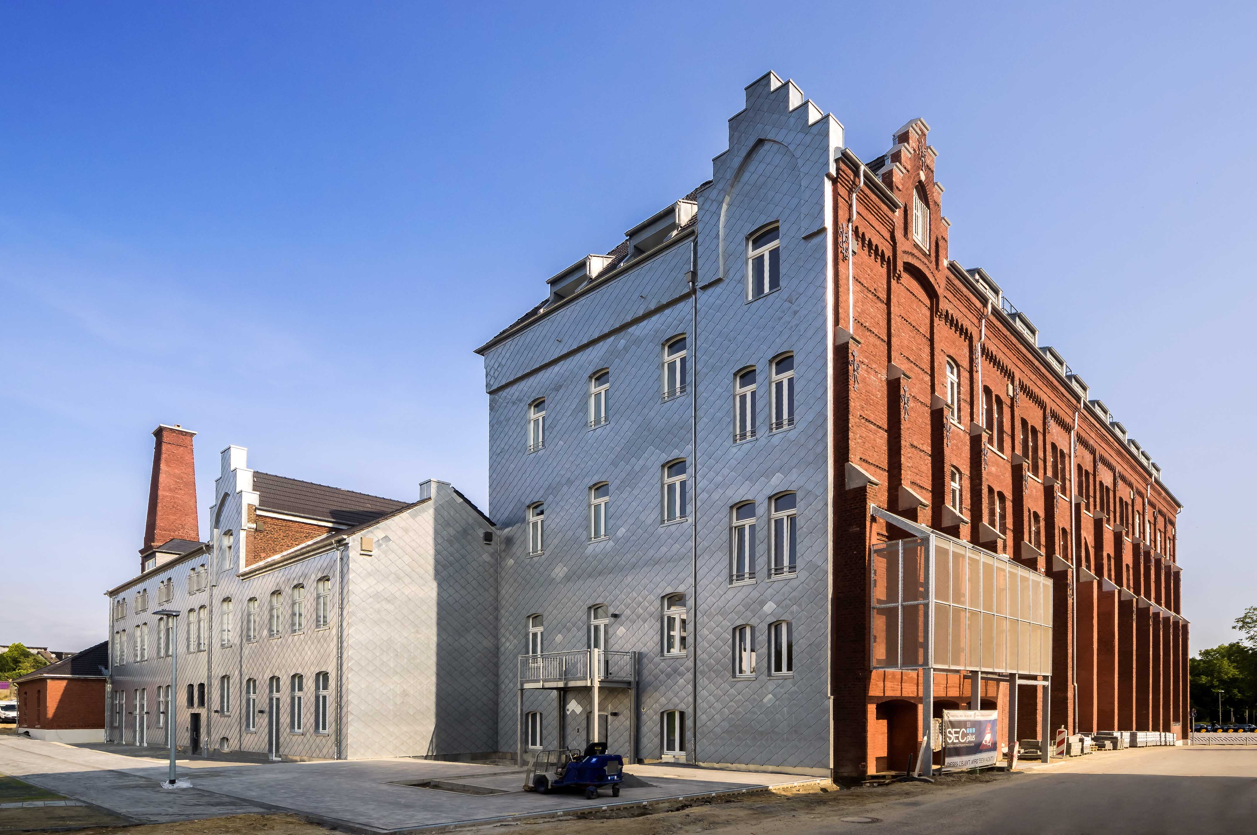 Außenansicht des Wohngebäudes Guter Hirte in Aachen mit roter Backsteinfassade und moderner Schieferverkleidung bei Tageslicht.