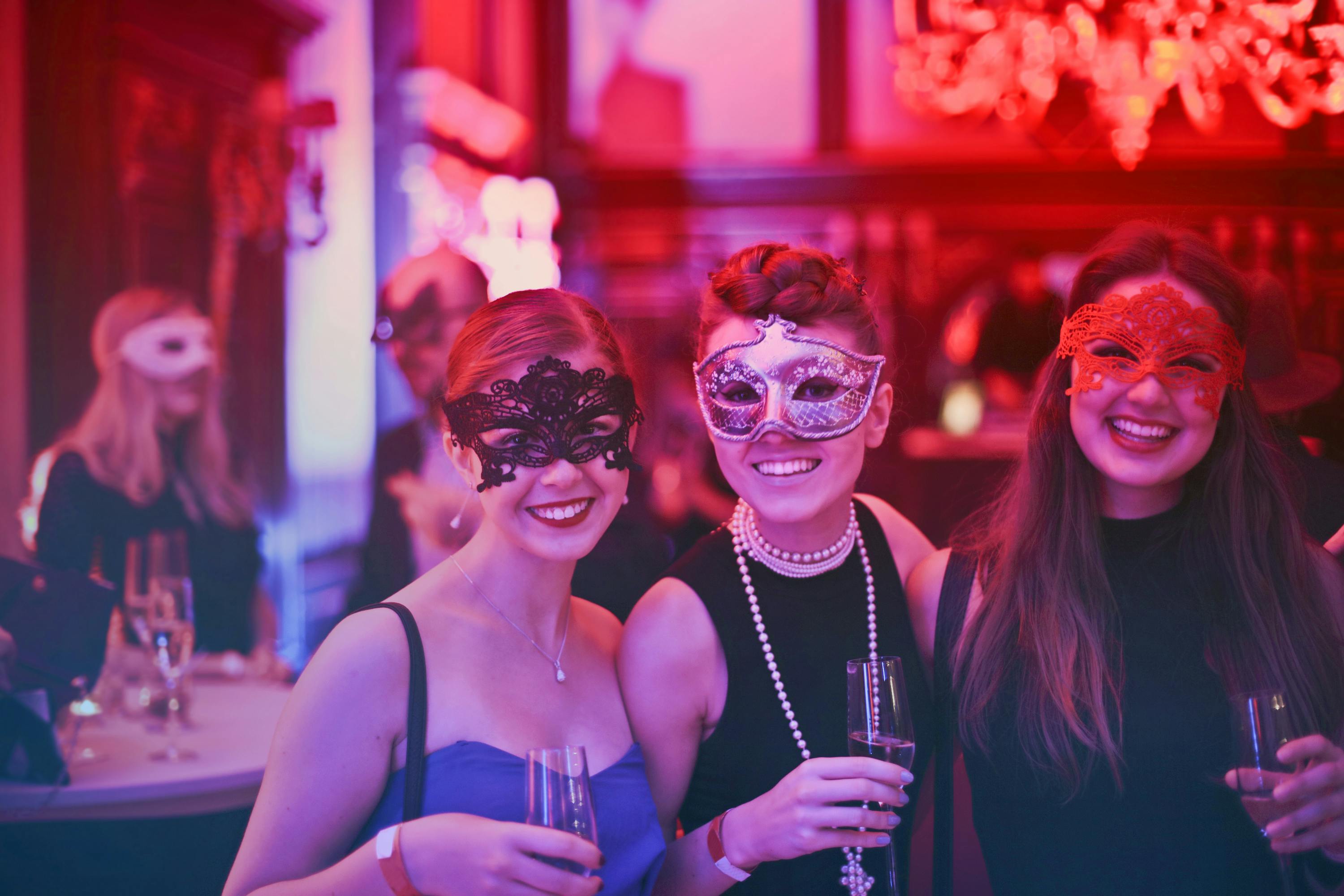Drei junge Frauen mit Masken auf einer festlichen Studentenparty in Aachen – fröhliche Stimmung, Abendgarderobe und Sektgläser in der Hand, Symbol für das lebendige Studentenleben der Stadt.