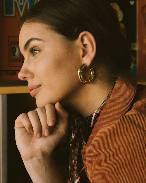 Mujer mirando hacia la izquierda, apoyando su mentón en la mano con expresión pensativa, lleva un pendiente dorado y una chaqueta marrón.