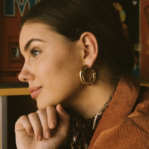 Mujer de perfil con pendientes grandes de aro y chaqueta marrón apoyando la barbilla en la mano.