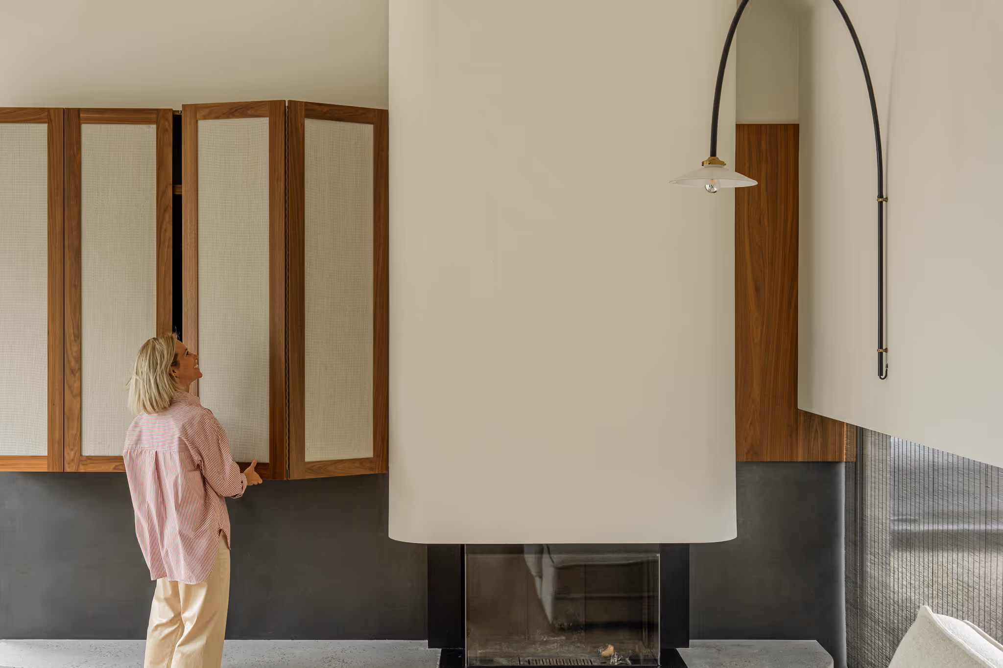 Moderne woonkamer met houten wandkasten en linnen deuren, vrouw in roze blouse opent kast naast minimalistische haard en design wandlamp.