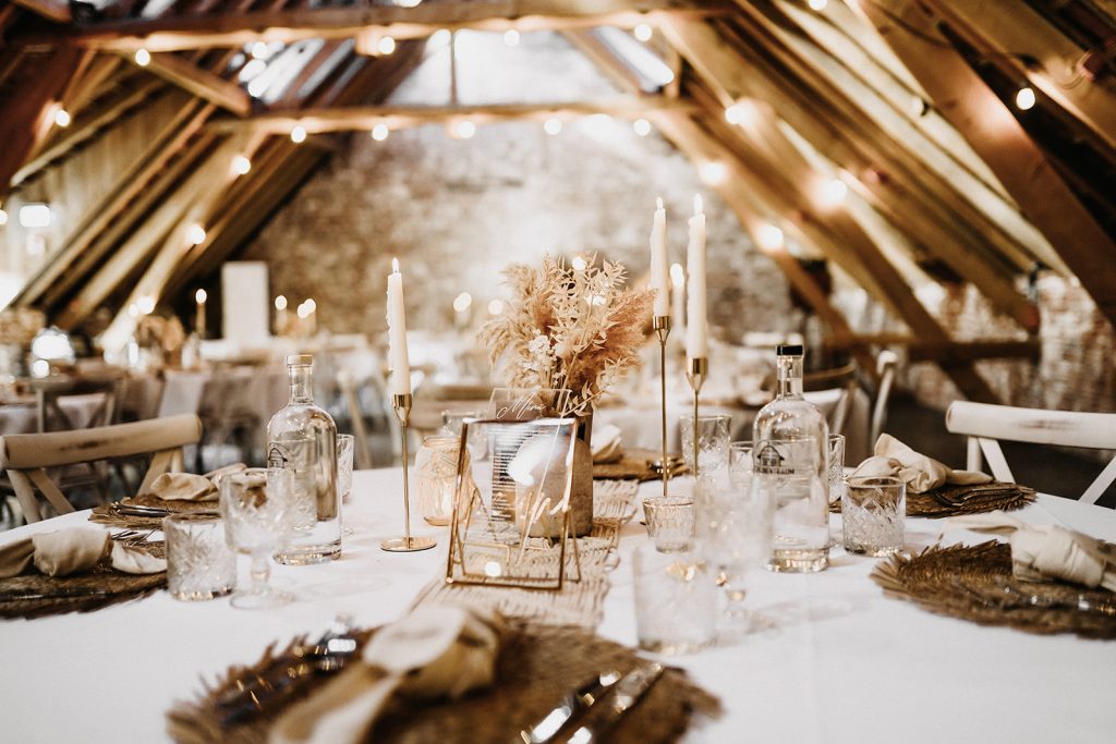 Rustikal dekorierter Festtisch mit weißen Kerzen, Glasflaschen und Trockenblumen unter Holzbalken in einem warm beleuchteten Raum.