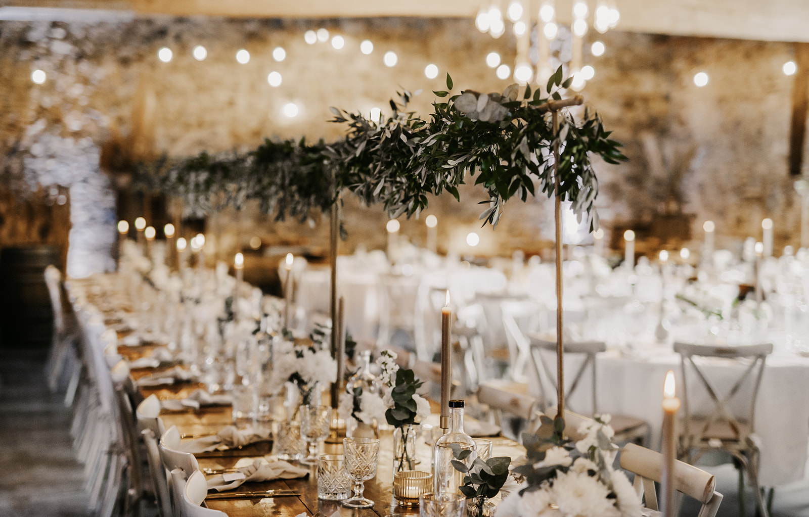 Eleganter festlich gedeckter Holztisch mit Kerzen, Glaswaren und Blumenarrangements in einem rustikalen Veranstaltungsraum.