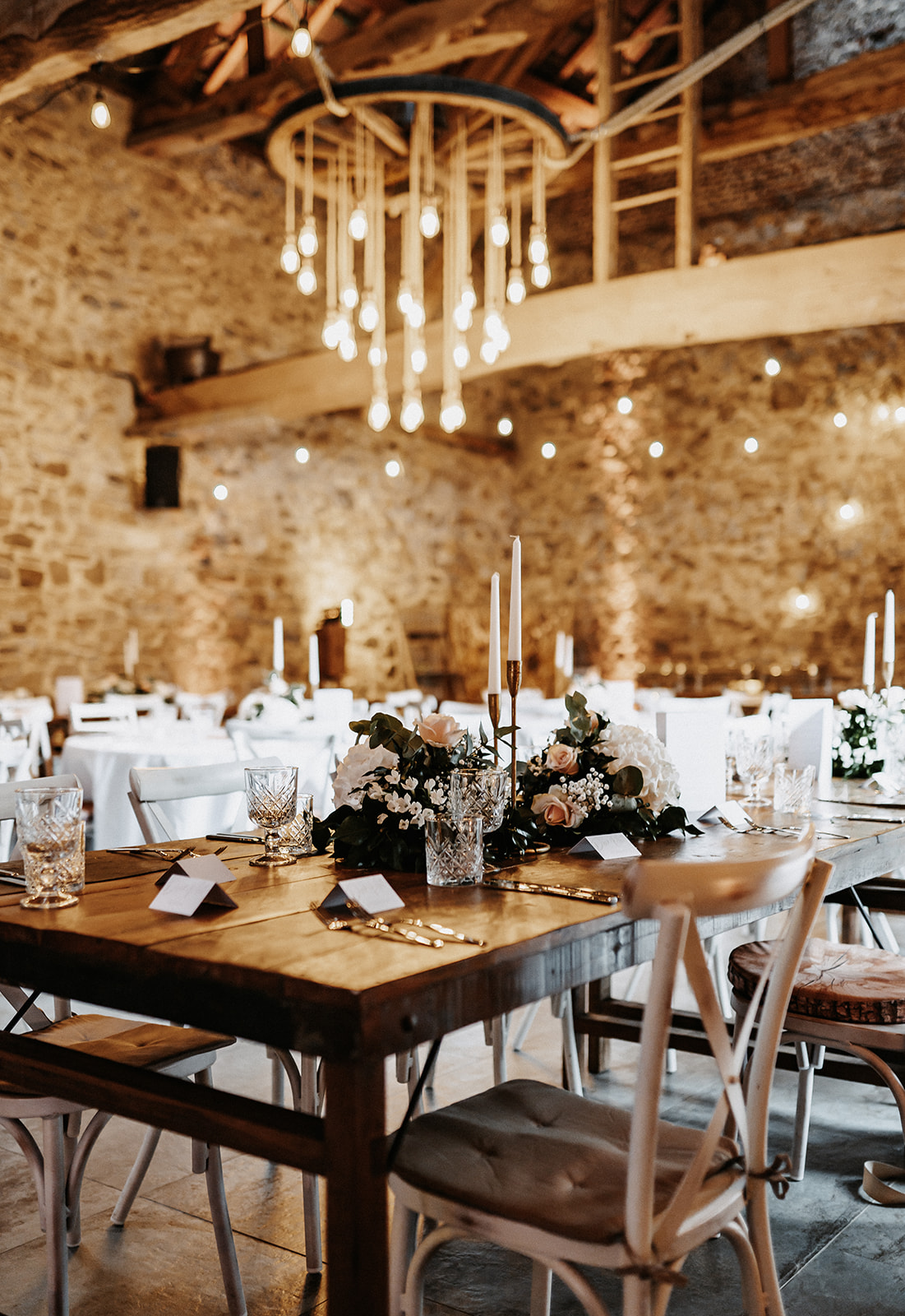 Rustikaler Festsaal mit Holztischen, weißen Stühlen, elegant dekorierten Tischarrangements und warmen Hängeleuchten.