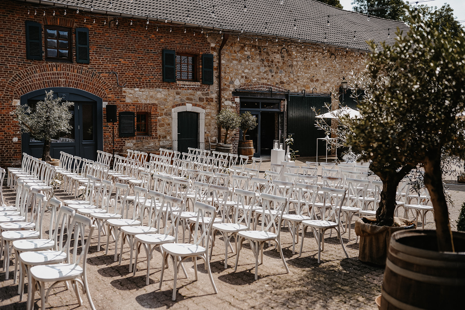 Draußen angeordnete weiße Stühle vor einem Backstein- und Steinbau mit grünen Fensterläden und Türen, dekoriert mit Olivenbäumen in Töpfen.
