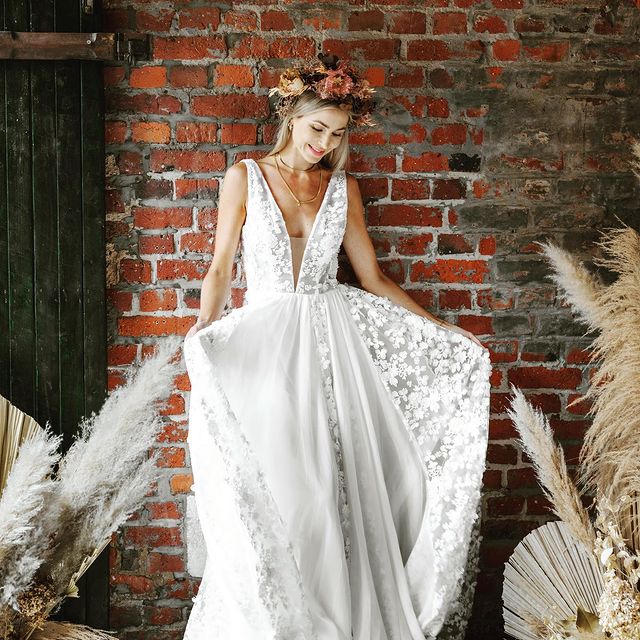 Frau in weißem Hochzeitskleid mit Blumenkopfschmuck vor roter Backsteinwand.