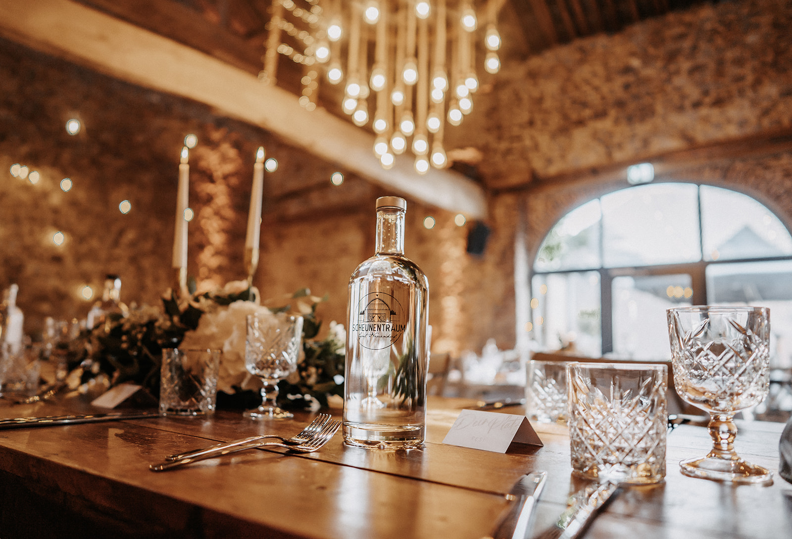 Elegantes Tischgedeck mit Glasflasche, Kristallgläsern und Kerzen in einem warm beleuchteten, rustikalen Raum mit Steinwänden.