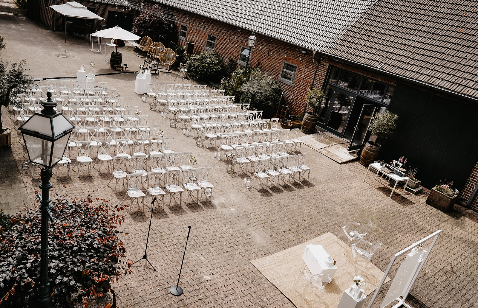 Außenbereich mit weißen Stühlen in Reihen vor einem Backsteingebäude, dekoriert für eine Zeremonie mit Mikrofonständern und Blumen.