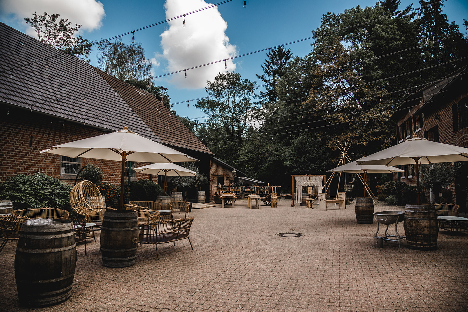 Innenhof mit Sitzgelegenheiten, Sonnenschirmen, Weinfässern und geschmücktem Holzrahmen vor Gebäuden aus rotem Backstein unter blauem Himmel.