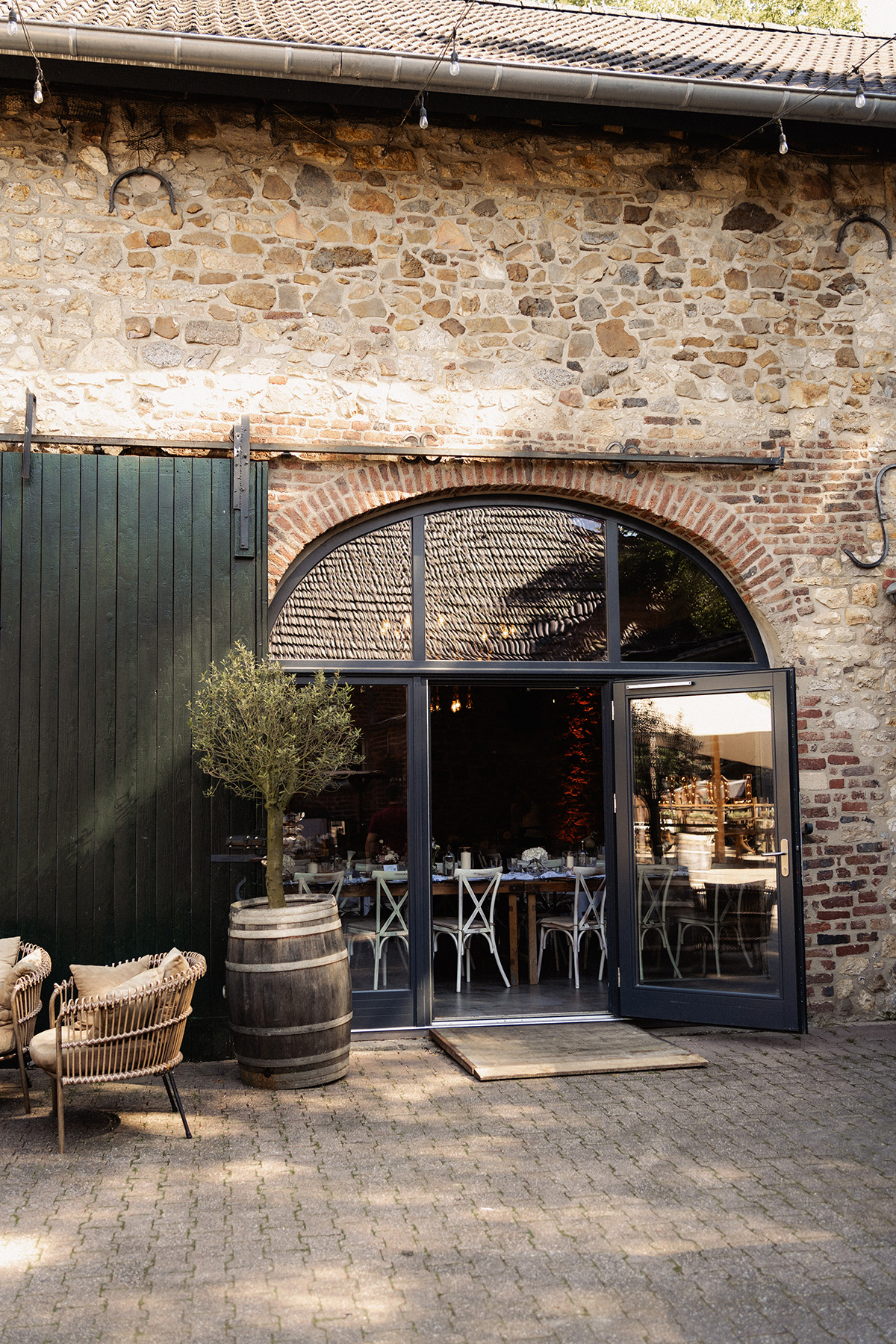 Eingang zu einem rustikalen Steingebäude mit Glastür, Holzbalken, Weinfass mit Baum und Korbsesseln draußen.