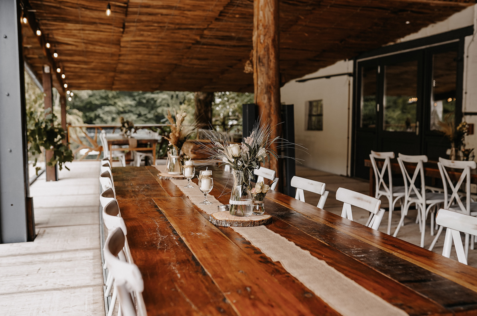 Rustikaler Holztisch mit weißer Tischdecke, dekoriert mit Blumen und Kerzen im überdachten Außenbereich mit weißen Stühlen.