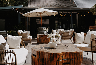 Gemütliche Außenterrasse mit hölzernen Tischen, weißen Sitzkissen, Stühlen und Sonnenschirm.