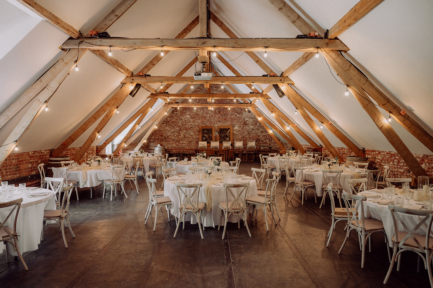 Hell beleuchteter Hochzeitsempfang in einem Dachboden mit Holzbalken und Ziegelwänden, rund gedeckte Tische mit weißer Tischdecke und Stühlen.
