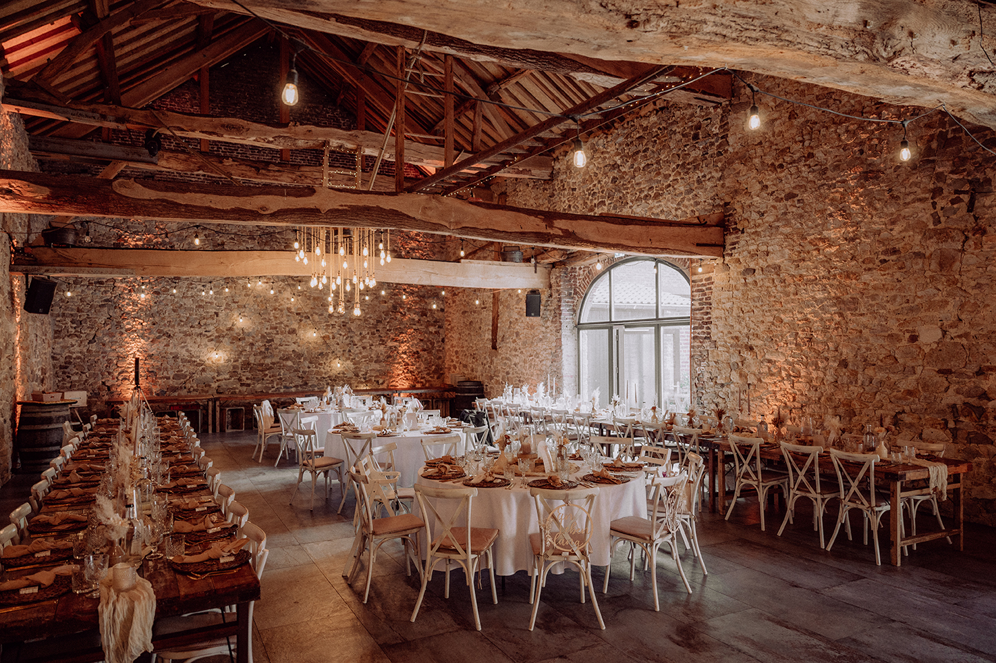 Rustikale festlich dekorierte Veranstaltungshalle mit Steinfassaden, Holzbalken und mehreren gedeckten Tischen mit weißen Stühlen.