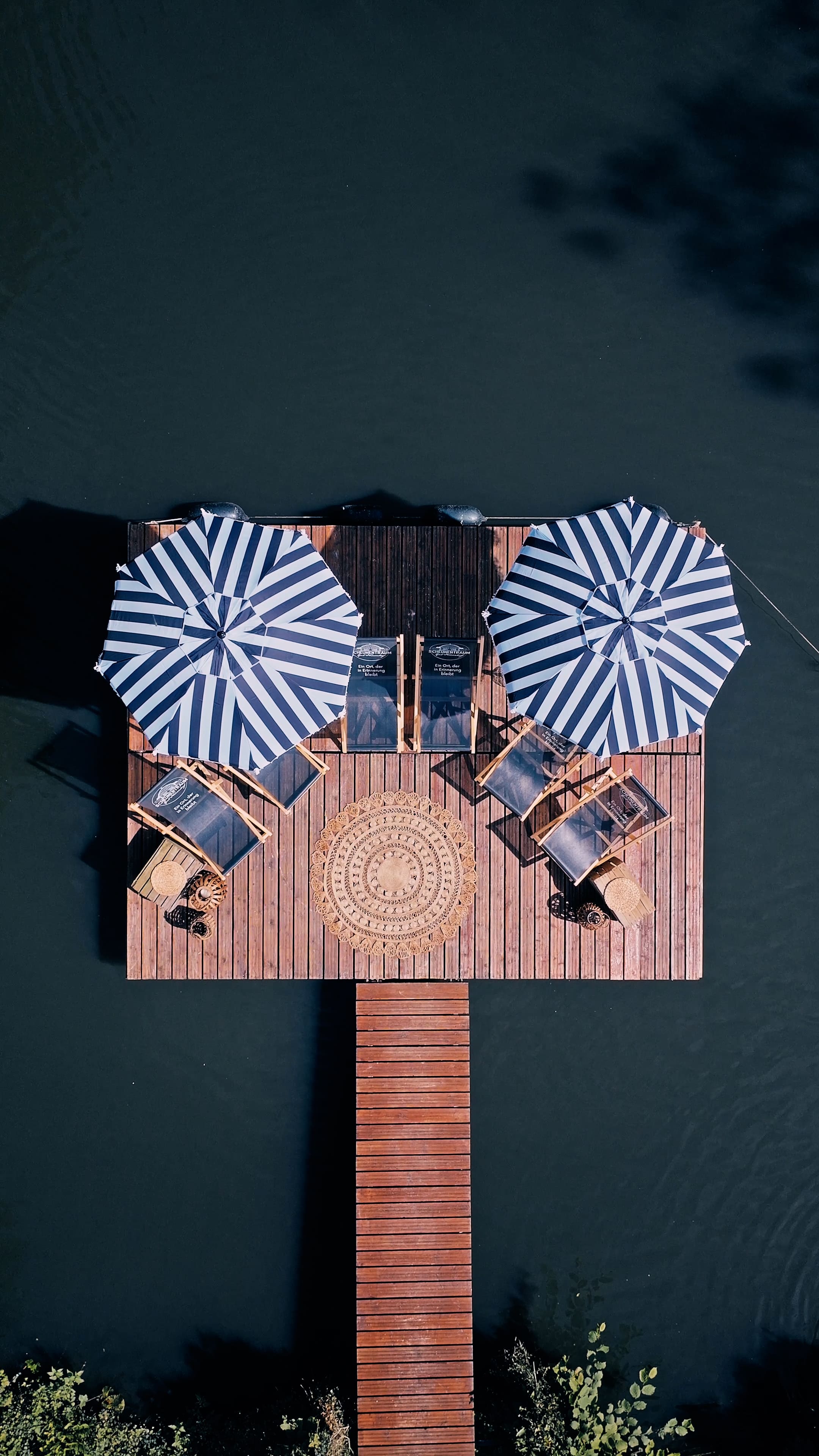 Luftaufnahme eines Stegs mit zwei gestreiften Sonnenschirmen, vier Liegestühlen und einem dekorativen runden Teppich darüber.