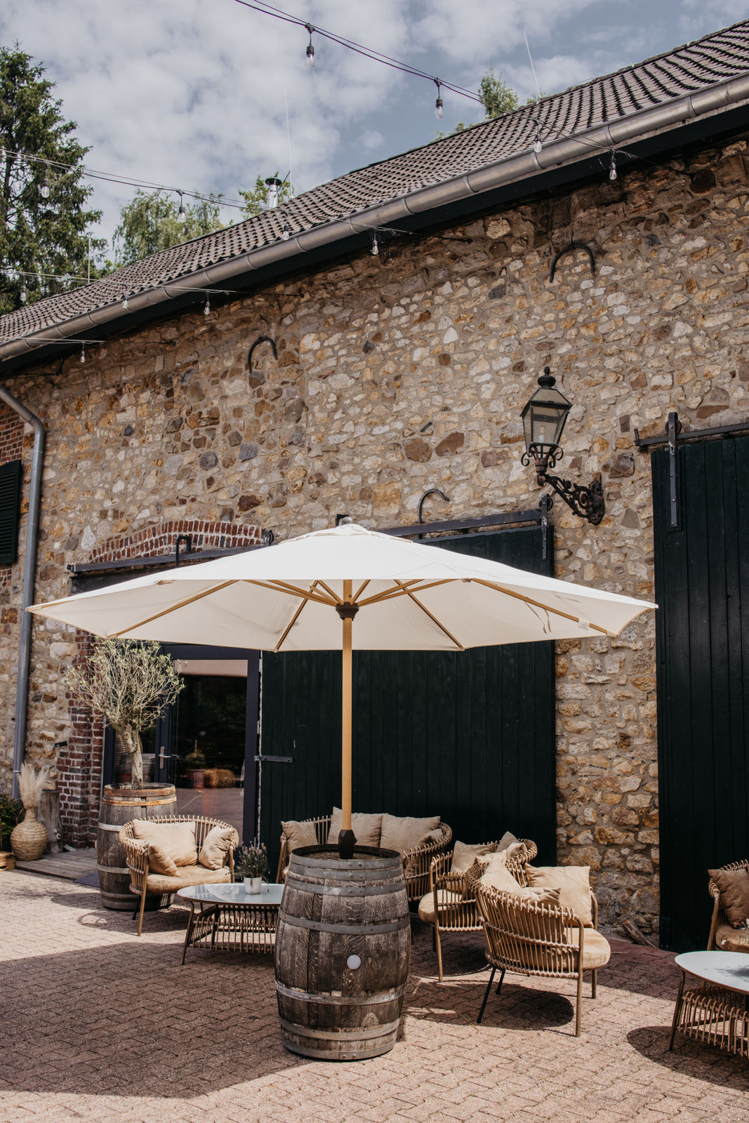 Gemütliche Außenbestuhlung mit Rattansesseln, einem Sonnenschirm auf einem Holzfass und Tischen vor einer Steinmauer mit schwarzen Türen.
