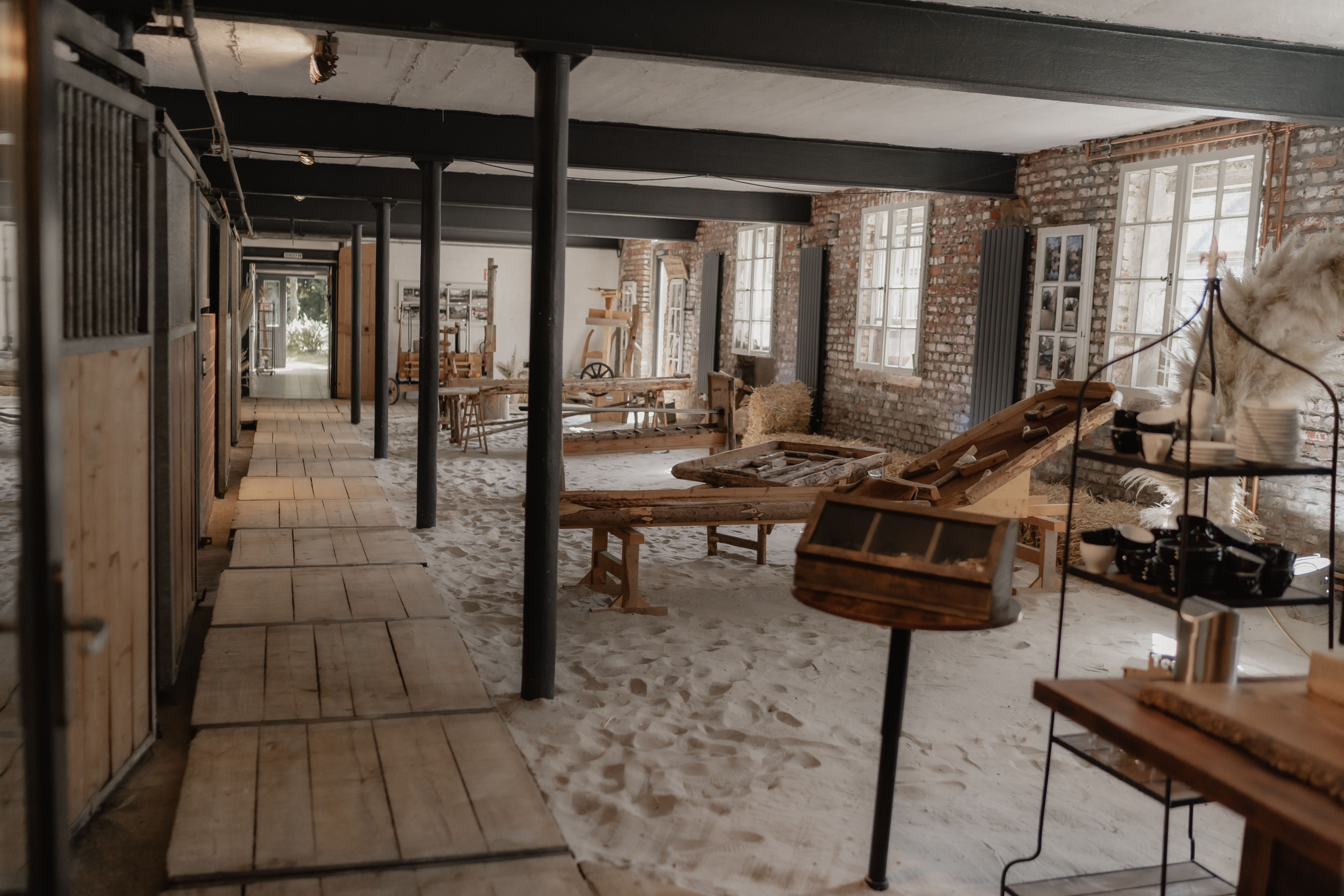 Innenansicht eines historischen, sandbedeckten Hofes mit Holzställen und landwirtschaftlichen Geräten.