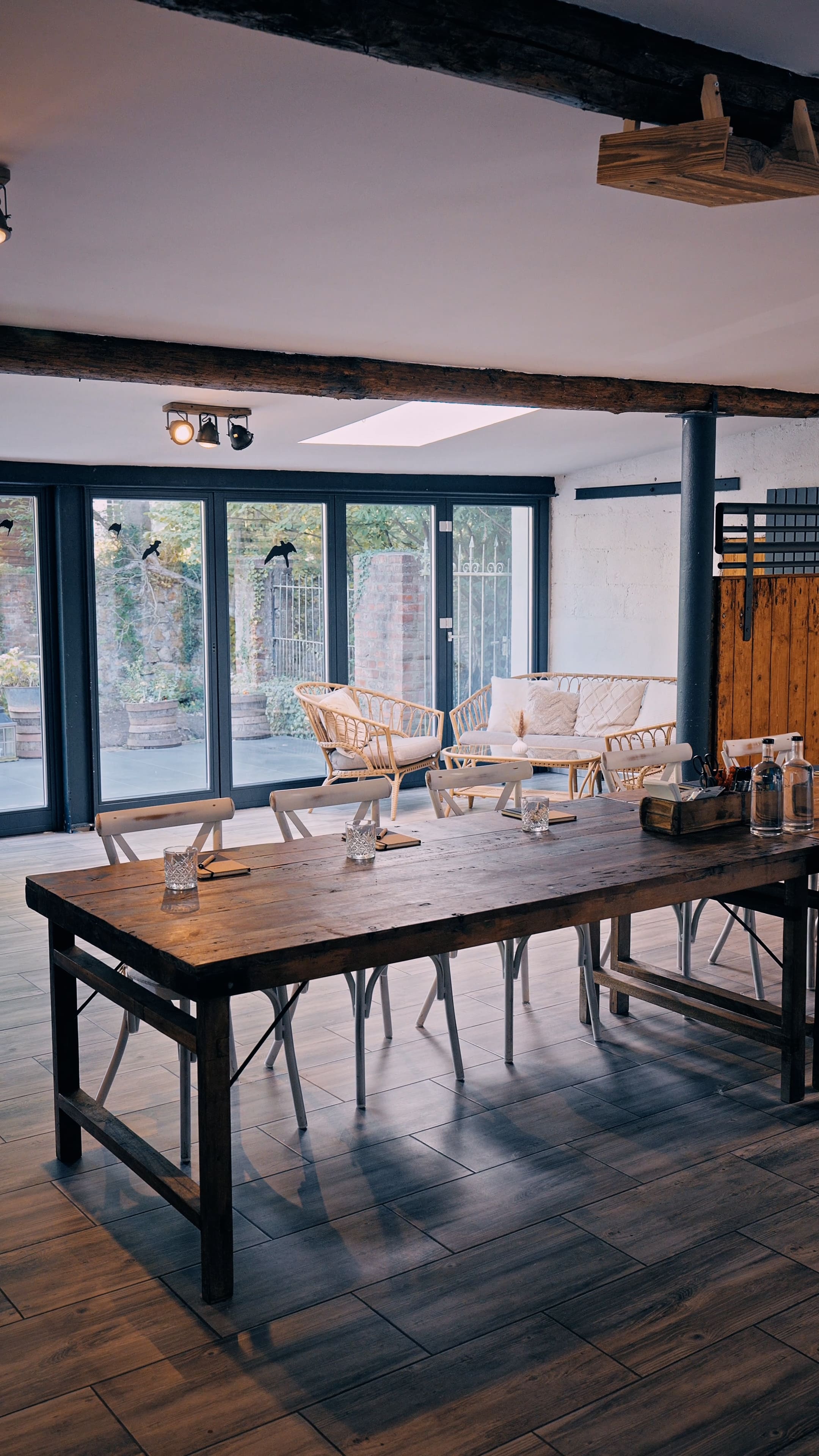 Rustikaler Holz-Tisch mit Stühlen in einem hellen Raum mit großen Fenstern und einem Sitzbereich mit Korbmöbeln.