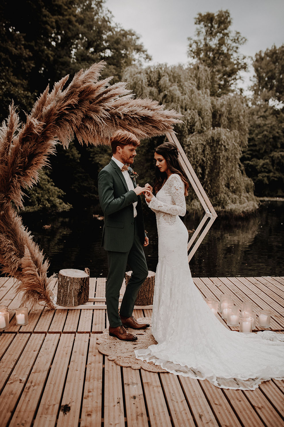 Braut und Bräutigam tauschen Ringe auf einem Holzsteg am See mit dekorativem Bogen aus Pampasgras und Kerzen.