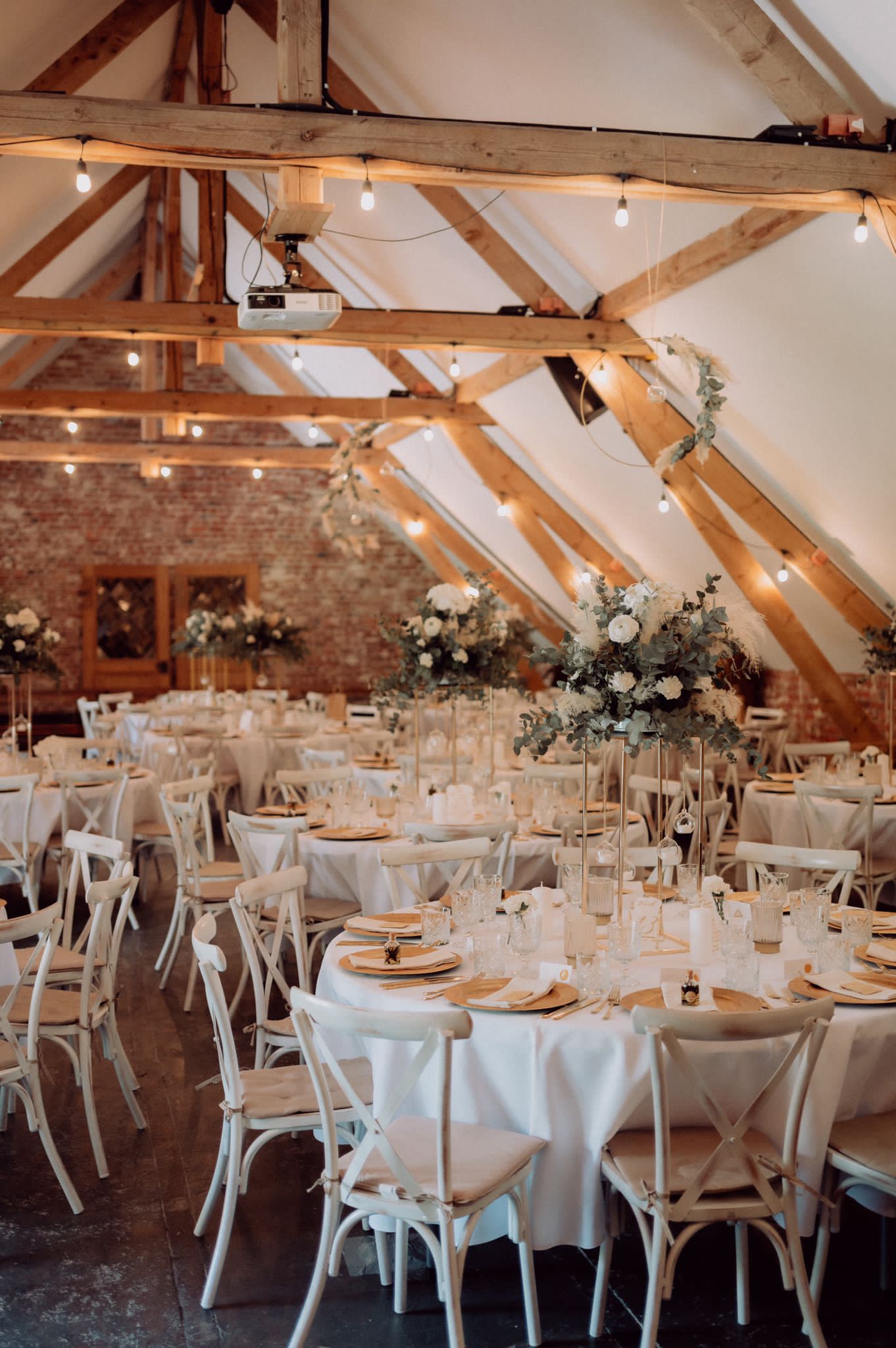 Eleganter Bankettsaal mit weißen Gartentischstühlen und rund gedeckten Tischen, geschmückt mit hohen Blumengestecken und warmen Lichterketten an Holzbalken.