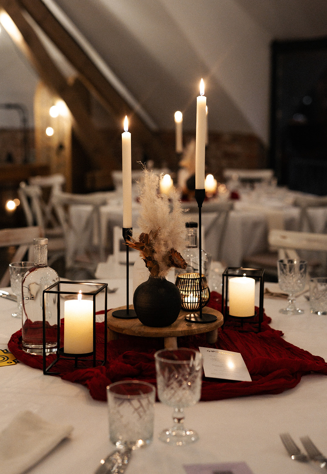 Festlich gedeckter Tisch mit brennenden Kerzen in Haltern, einer Vase mit getrockneten Blumen und Kristallgläsern auf weißem Tischtuch mit rotem Stoff.