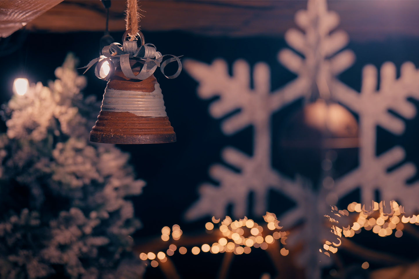 Nahaufnahme einer hängenden glasierten Keramikglocke vor verschwommenen Weihnachtsbeleuchtungen und einer großen Schneeflockendekoration.