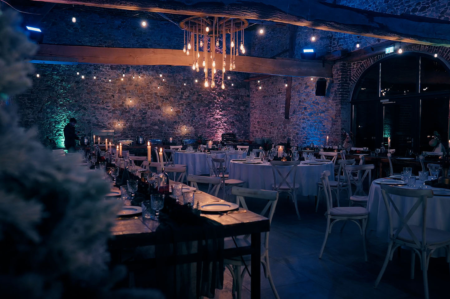Gemütlich beleuchteter Festsaal mit rustikalen Steintischen, weißen Stühlen, runden gedeckten Tischen, Kerzen und Hängelampen.