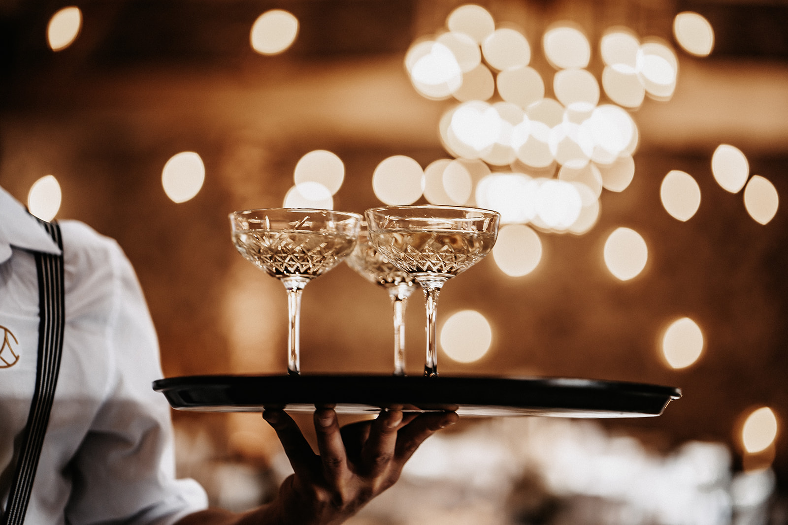 Kellner hält ein Tablett mit drei gefüllten Kristallsektschalen vor einem verschwommenen, warm beleuchteten Hintergrund.