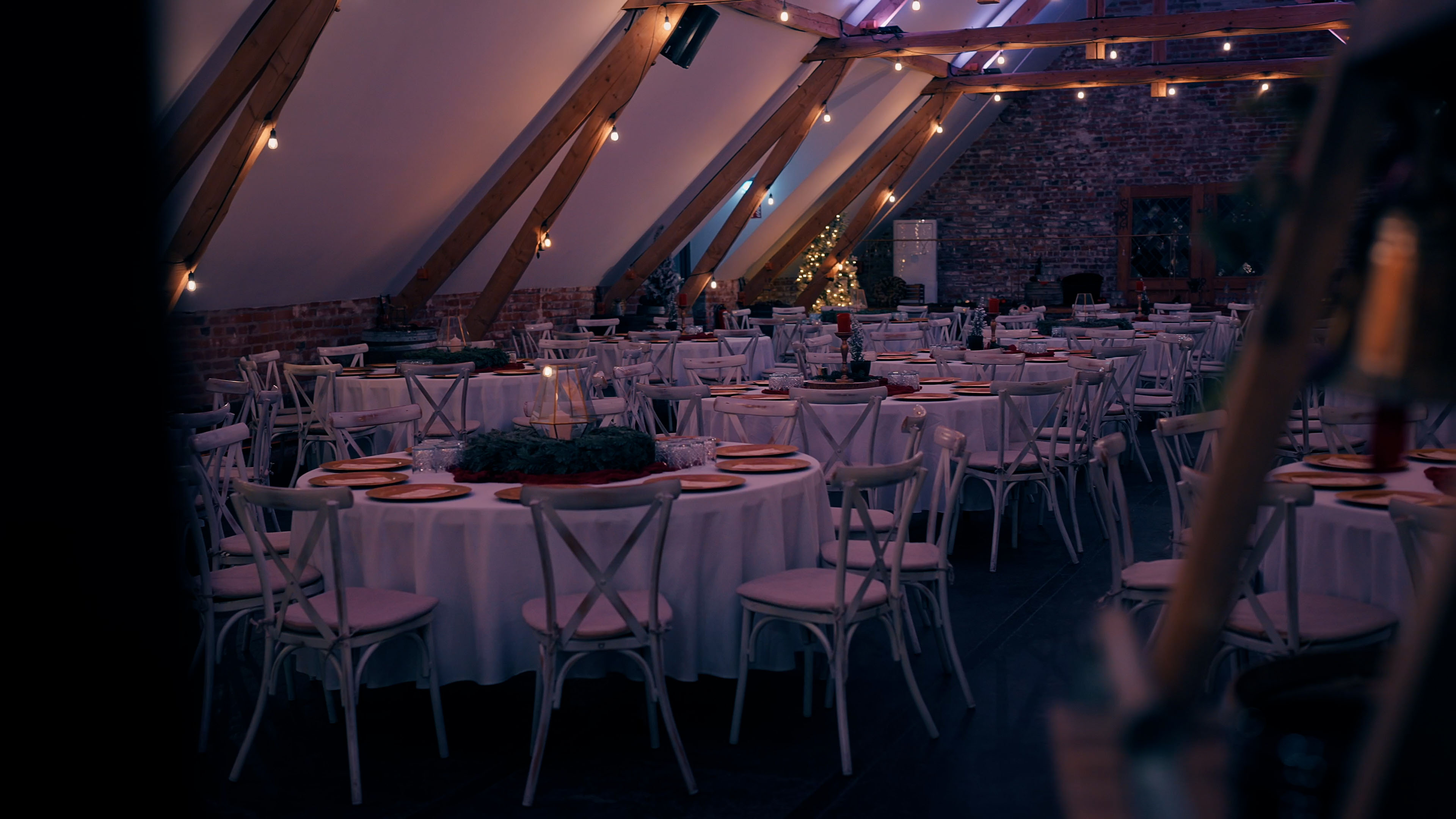 Weihnachtlich dekorierter Saal mit runden Tischen, weißen Stühlen, Kerzen und leuchtenden Girlanden unter einer Dachschräge.