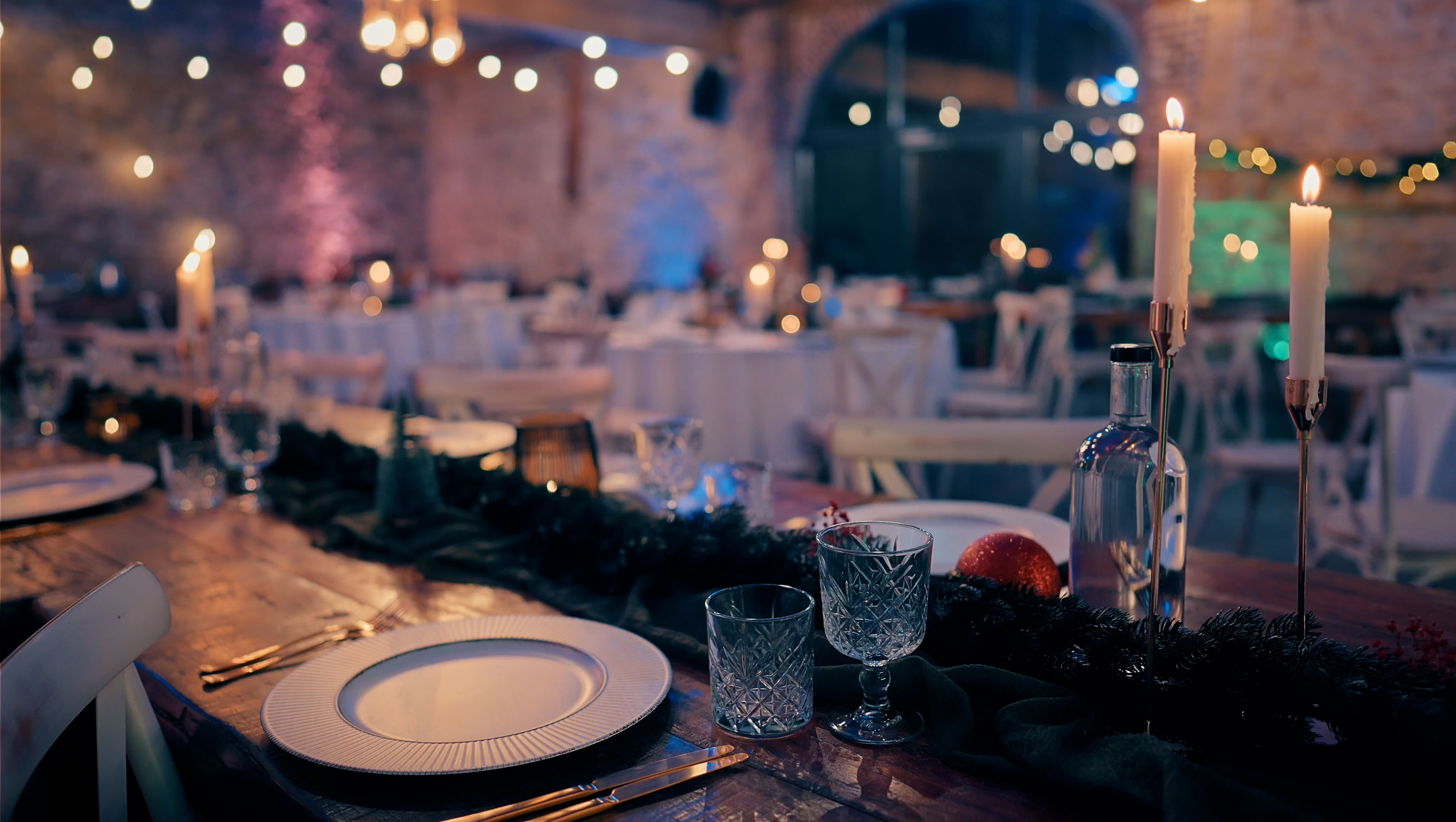 Festlich gedeckter Holztisch mit Kerzen, Glasflasche, Tannenzweigen und Weihnachtskugel in warm beleuchtetem Raum.