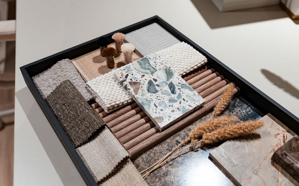 Flat lay of interior design material samples including stone, terrazzo, wood, fabric, and dried grasses.