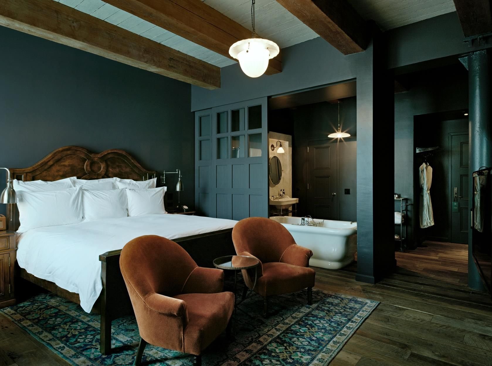 Moody luxury bedroom with exposed beams, freestanding bath, velvet armchairs, dark paneled walls