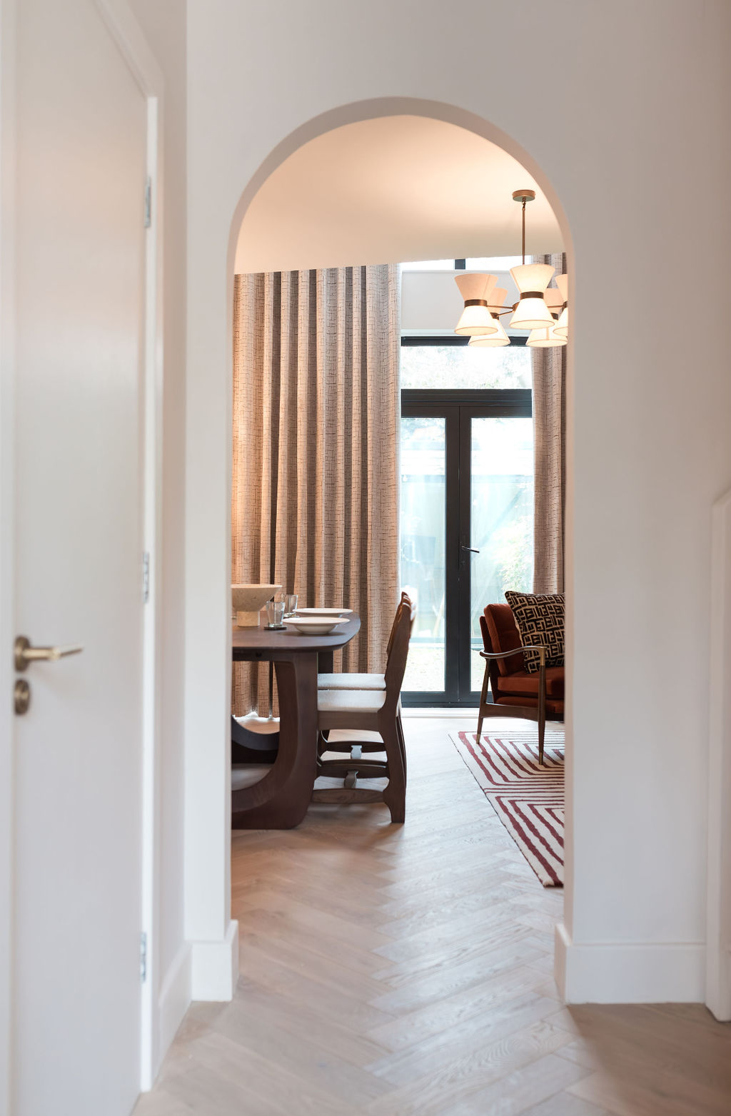 View through an arched doorway in an Islington townhouse by TXTURED, showing a dining area with warm timber furniture, soft neutral curtains, layered lighting and earthy accents beyond.