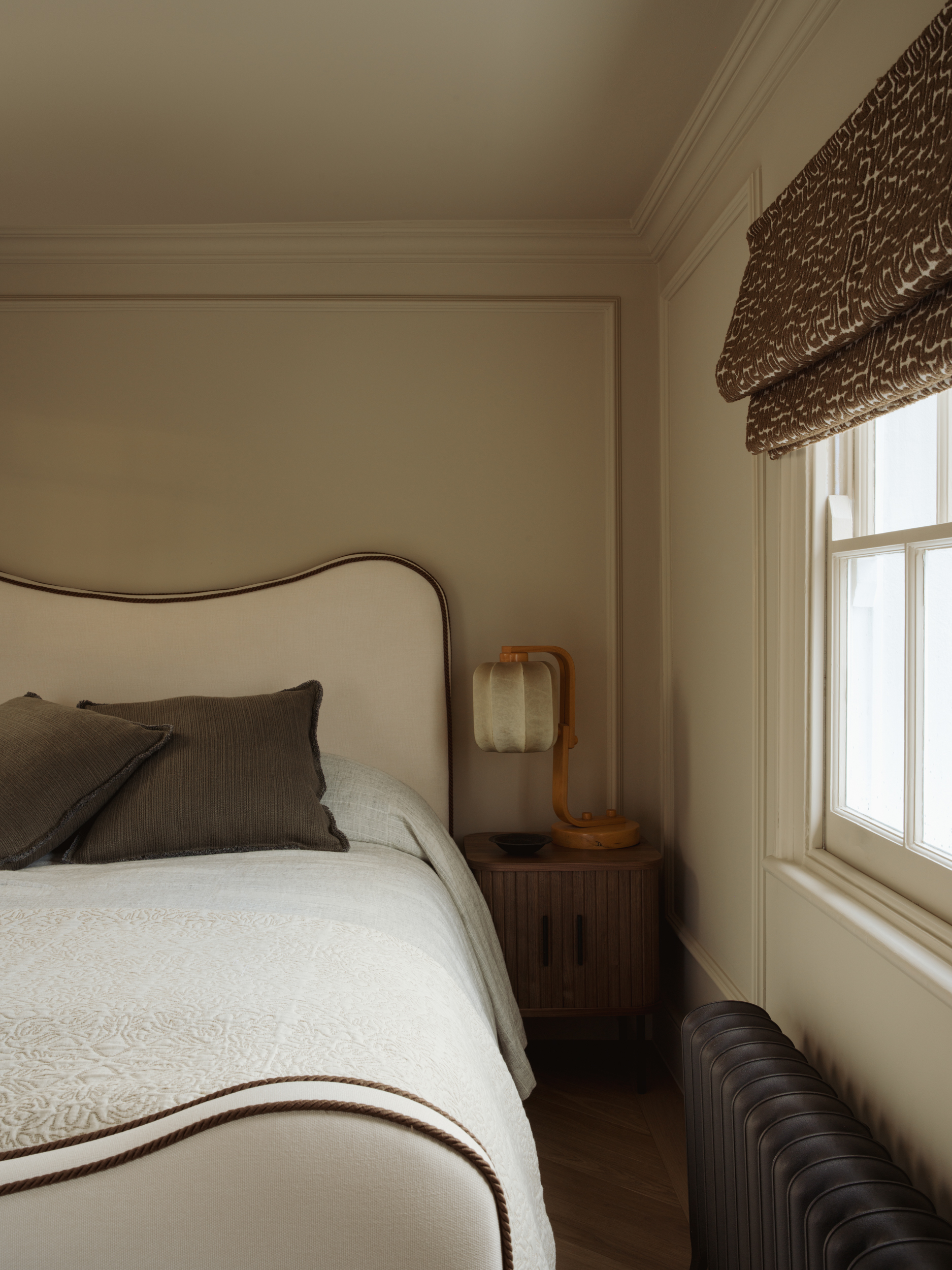 A beautifully styled bedroom corner featuring an upholstered bedhead with curved edges and contrast piping, layered with olive linen cushions and a cream quilted bedspread. A fluted walnut bedside cabinet sits beside the bed, topped with a sculptural amber table lamp. A dark cast iron radiator and white sash window complete the scheme.