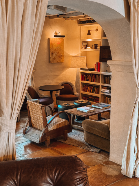A warm, intimate lounge area at Soho House Ibiza, framed by a white arched doorway and linen curtains, with a mix of vintage wooden and velvet armchairs, a low wooden coffee table, and a built-in bookshelf lit by warm wall sconces.