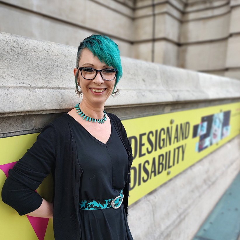 This is me! I've got an asymmetrical turquoise fringe with a matching necklace and belt with a black dress, cardigan, glasses and spiky earrings. I'm standing in front of a banner advertising the Design and Disability exhibition at the V&A in London.