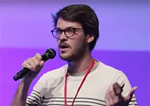 Un homme portant des lunettes et un t-shirt clair parle dans un microphone sur une scène avec un fond violet.